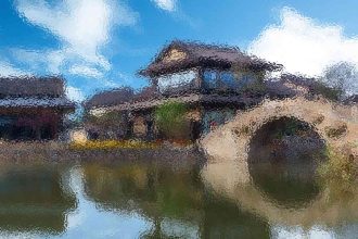 House with a large circular water wheel in front, beside a calm pond under a blue sky with clouds.