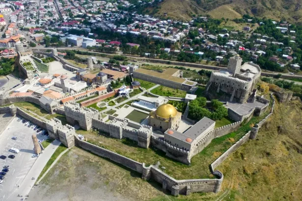 Aerial view of a medieval fortress with tall stone walls and towers, a gold-domed building in a courtyard, and a town below on surrounding hillsides.