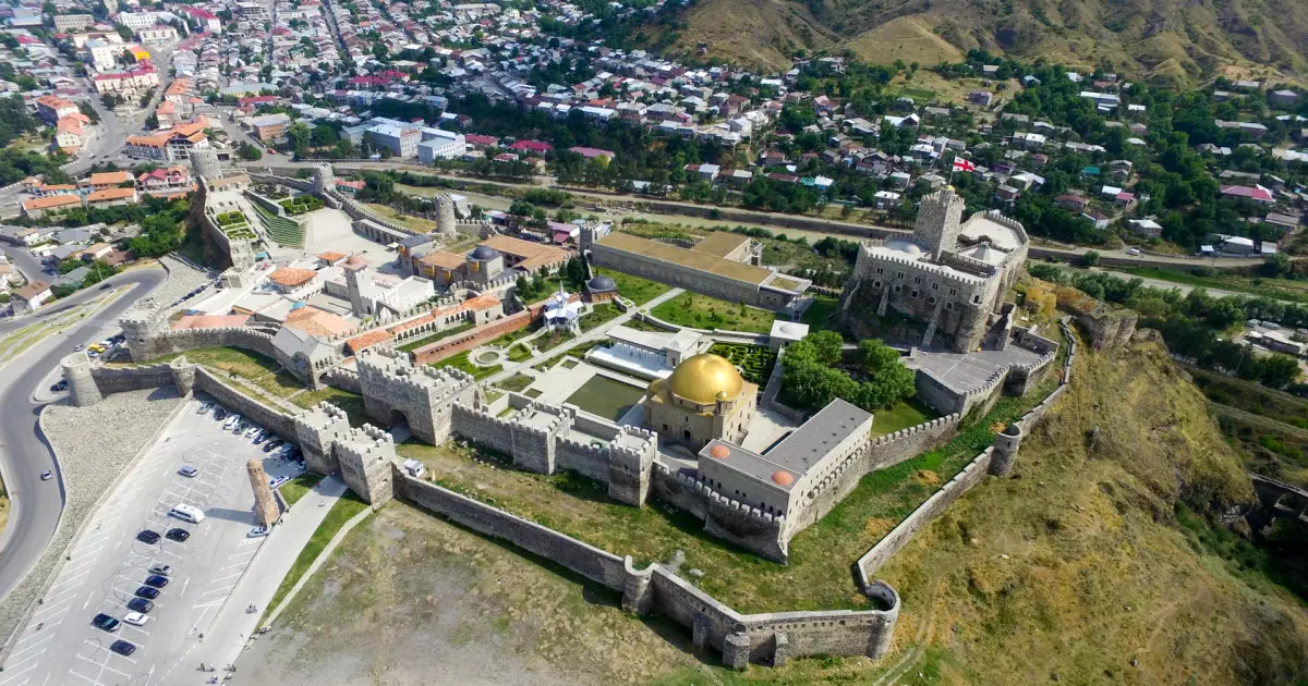 Aerial view of a medieval fortress with tall stone walls and towers, a gold-domed building in a courtyard, and a town below on surrounding hillsides.