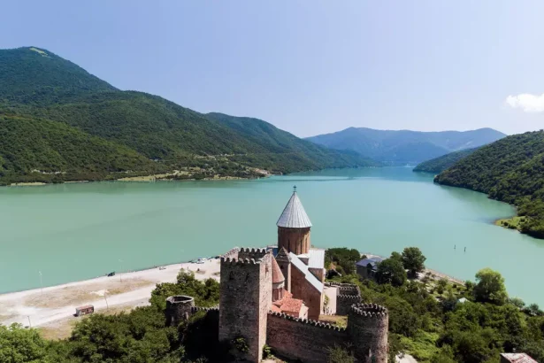 Stone fortress with a cylindrical tower overlooks a turquoise lake and forested mountains. (Aerial view)