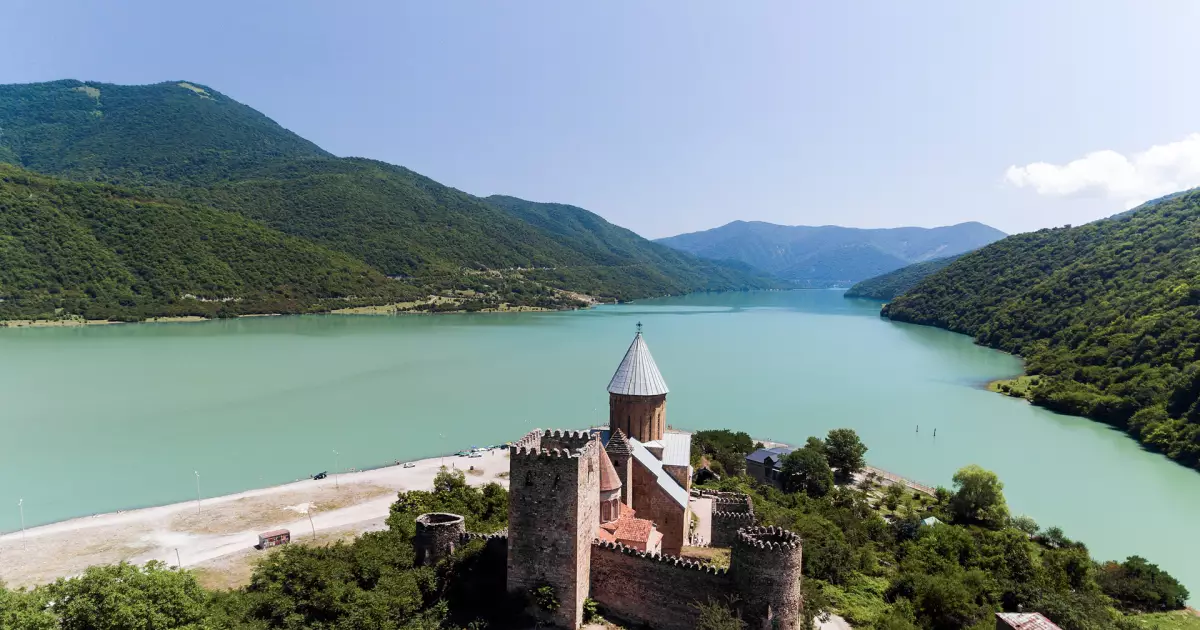 Stone fortress with a cylindrical tower overlooks a turquoise lake and forested mountains. (Aerial view)