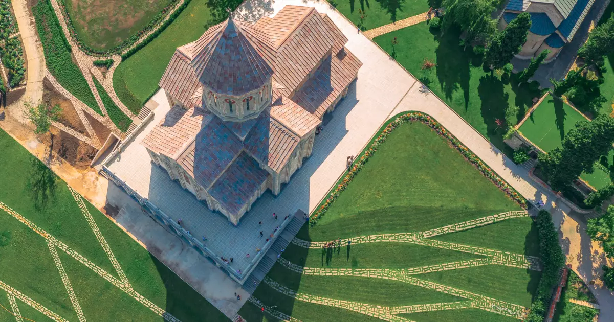 Aerial view of a stone church with a copper roof, surrounded by manicured lawns, flower beds, and intersecting stone pathways.
