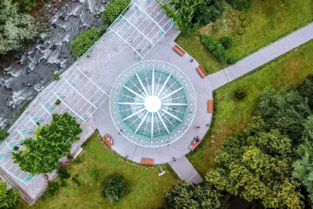 Aerial view of a circular glass-roofed pavilion in a green park, with benches, paths, and surrounding trees.