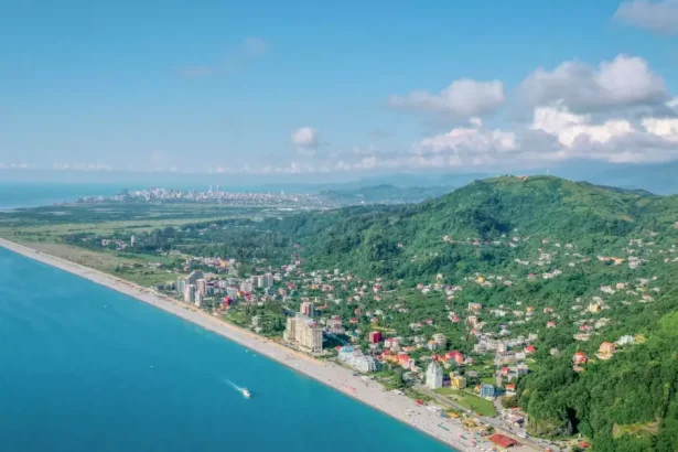 Aerial view of a seaside town with a long sandy beach, turquoise water, and green hills dotted with houses inland.