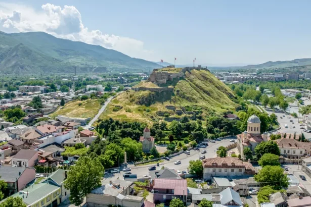 Aerial view of a hilltop fortress with flags overlooking a town with red-roofed houses and a domed church below.