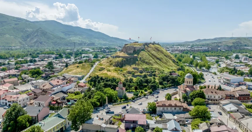 Aerial view of a hilltop fortress with flags overlooking a town with red-roofed houses and a domed church below.
