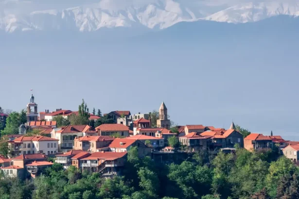 A hillside town with red-tiled roofs nestled among trees under a pale blue sky and distant mountains.