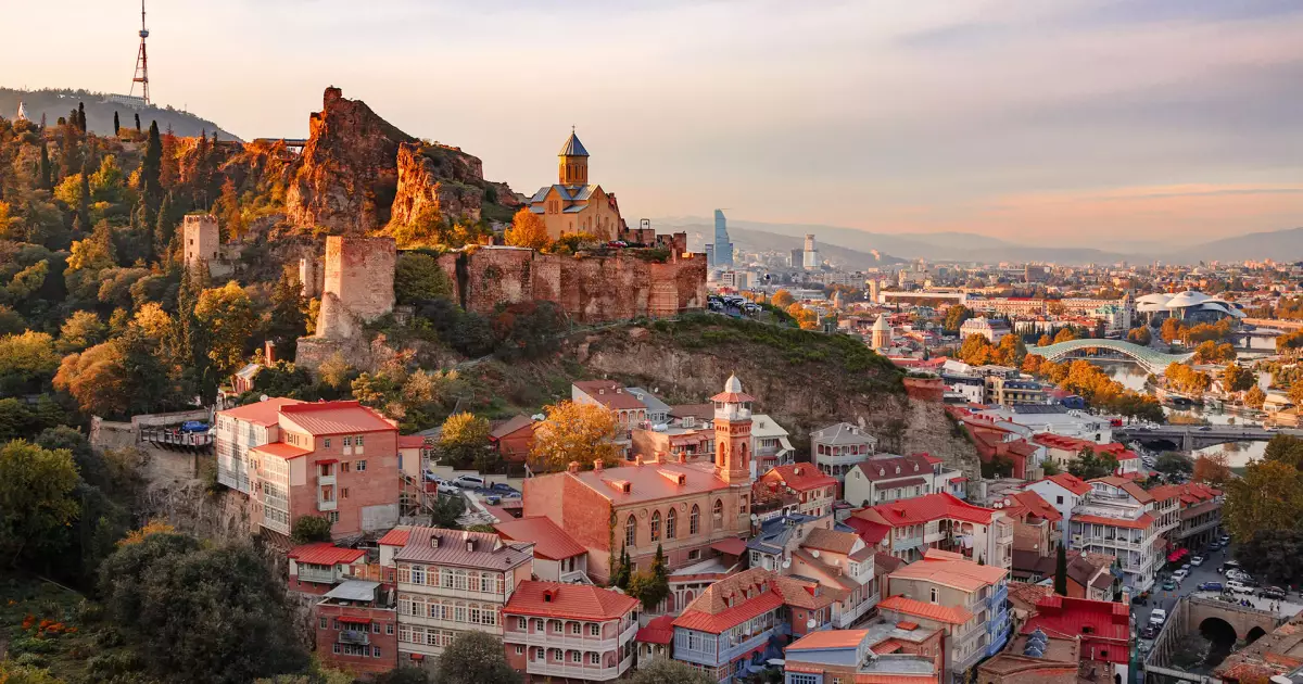 Aerial view of Tbilisi's old town with Narikala Fortress on a hill, red-roofed houses cascading down the slope at sunset.