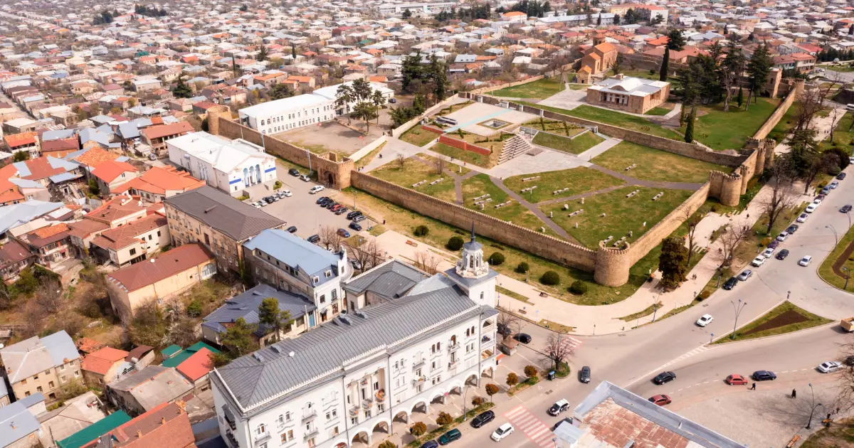 Aerial view of a historic fortress with fortified walls surrounding a green park and city buildings nearby, with a large white government-style building at the corner street.
