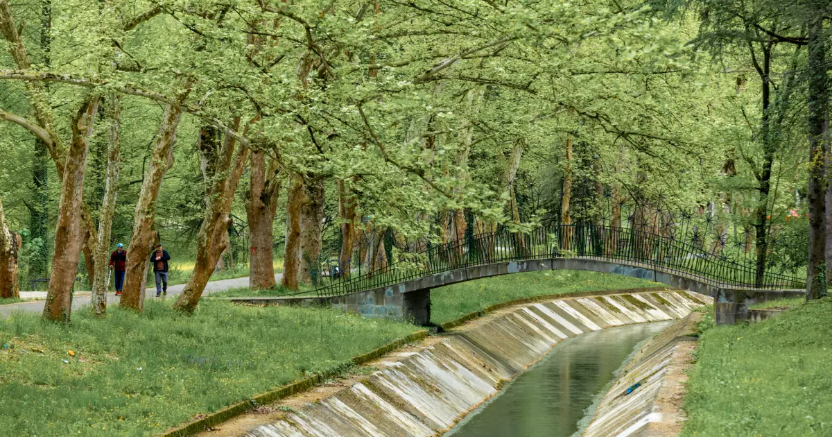 Park scene with a canal and a curved metal bridge among leafy trees; two people walk along the path in the distance.