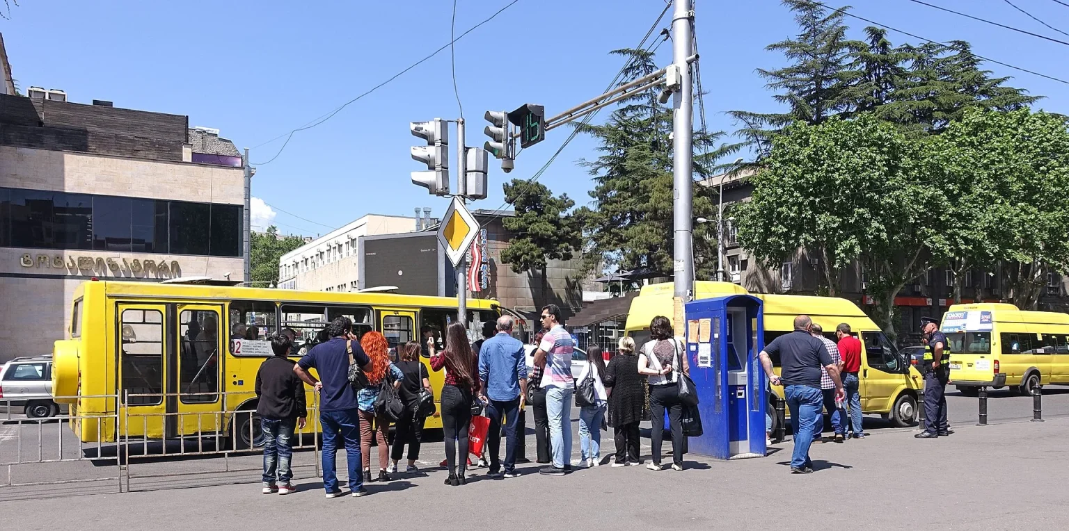 Passengers board a yellow city bus at a busy Tbilisi bus stop on Merab Kostava Street.