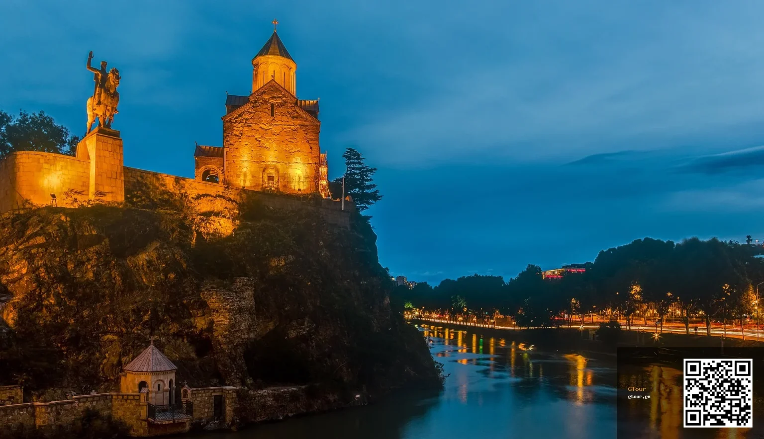 Metekhi Church and the Kartlis Deda statue overlook the Kura River in Tbilisi's old town at dusk.