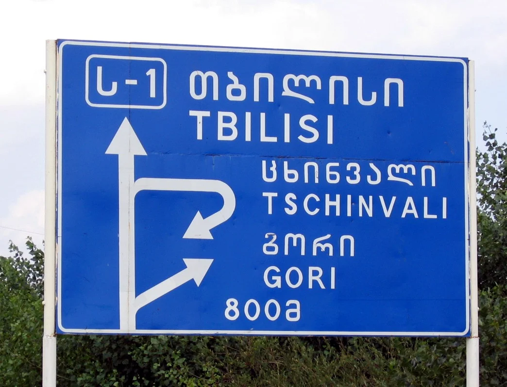 A road sign near Tbilisi shows place names in both Georgian Mkhedruli script and Latin transliteration.